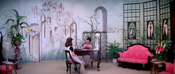Movie still from “Suspiria” (1977), directed by Dario Argento – Two women sitting at a table in front of a painting; Wide shot, High angle