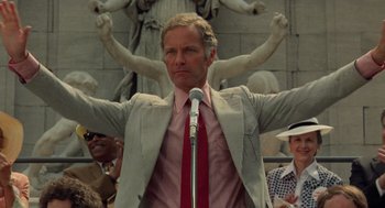 Movie still from “Taxi Driver” (1976), directed by Martin Scorsese – A man in a suit and red neck tie holding a microphone in front of a crowd; Medium shot, Low angle