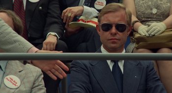Movie still from “Taxi Driver” (1976), directed by Martin Scorsese – A man in a suit and sunglasses sitting next to a crowd; Close Up shot, Over the shoulder angle