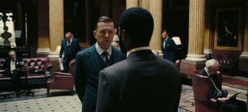 Movie still from “Tenet” (2020), directed by Christopher Nolan – A man in a suit and tie standing next to another man in a suit and tie; Medium shot, Over the shoulder angle