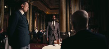 Movie still from “Tenet” (2020), directed by Christopher Nolan – A man in a suit standing in front of a table; Medium shot, Over the shoulder angle