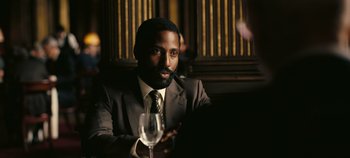 Movie still from “Tenet” (2020), directed by Christopher Nolan – A man sitting at a table holding a wine glass; Close Up shot, Over the shoulder angle