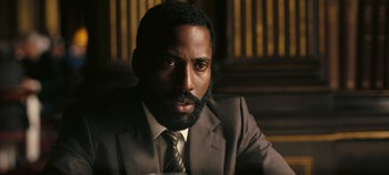 Movie still from “Tenet” (2020), directed by Christopher Nolan – A man wearing a suit and tie with a goatee; Close Up shot, Over the shoulder angle