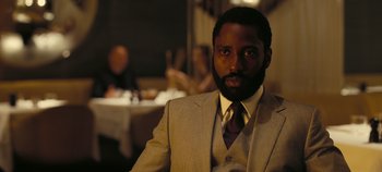 Movie still from “Tenet” (2020), directed by Christopher Nolan – A man wearing a suit and tie sitting in front of a table; Close Up shot, Over the shoulder angle