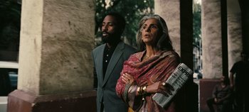Movie still from “Tenet” (2020), directed by Christopher Nolan – A man standing next to an older woman holding a newspaper; Medium shot, Low angle