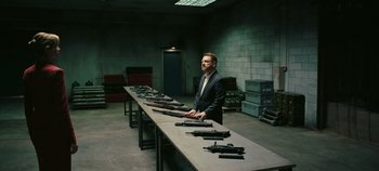 Movie still from “Tenet” (2020), directed by Christopher Nolan – A man standing in front of a long table of guns; Wide shot, High angle