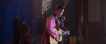 Movie still from “Elvis” (2022), directed by Baz Luhrmann – A man in a pink jacket holding a guitar and a microphone; Medium shot, Over the shoulder angle