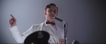 Movie still from “Elvis” (2022), directed by Baz Luhrmann – A man in a white suit and red bowtie is holding a microphone; Close Up shot, Low angle