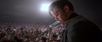 Movie still from “Elvis” (2022), directed by Baz Luhrmann – A man is holding a microphone in front of a crowd; Medium shot, Over the shoulder angle