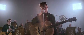 Movie still from “Elvis” (2022), directed by Baz Luhrmann – A man holding a microphone and holding a guitar; Medium shot, Over the shoulder angle