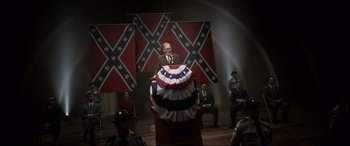 Movie still from “Elvis” (2022), directed by Baz Luhrmann – A man in a suit and tie standing in front of an american flag; Wide shot, Low angle
