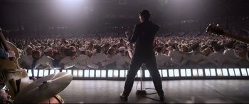 Movie still from “Elvis” (2022), directed by Baz Luhrmann – A man in a black suit is on a stage with a microphone; Wide shot, High angle