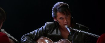 Movie still from “Elvis” (2022), directed by Baz Luhrmann – A man in a black jacket is playing a guitar; Close Up shot, Over the shoulder angle