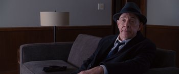 Movie still from “The Accountant” (2016), directed by Gavin O'Connor – An older man sitting on a couch wearing a suit and hat; Medium shot, Over the shoulder angle