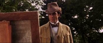 Movie still from “The Age of Innocence” (1993), directed by Martin Scorsese – A man wearing a hat and a bow tie standing in front of a wooden structure; Close Up shot, Over the shoulder angle
