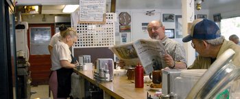 Movie still from “The Big Short” (2015), directed by Adam McKay – A man sitting at a counter reading a newspaper; Medium shot, Over the shoulder angle