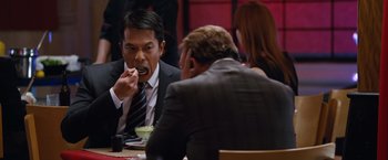 Movie still from “The Big Short” (2015), directed by Adam McKay – A man in a business suit eating a piece of food; Medium shot, Over the shoulder angle