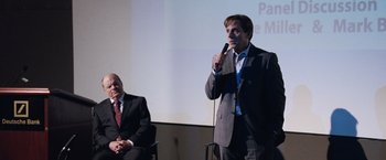 Movie still from “The Big Short” (2015), directed by Adam McKay – A man standing at a microphone with another man sitting in front of him; Medium shot, Over the shoulder angle