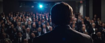 Movie still from “The Big Short” (2015), directed by Adam McKay – A crowd of people in a room with a man in a black suit; Medium shot, Over the shoulder angle