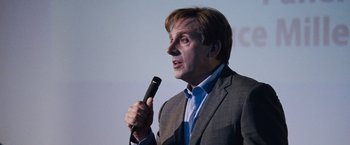 Movie still from “The Big Short” (2015), directed by Adam McKay – A man in a gray suit is holding a microphone; Close Up shot, Over the shoulder angle