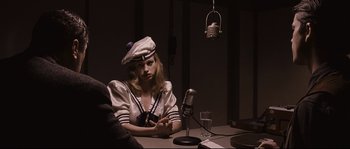 Movie still from “The Black Dahlia” (2006), directed by Brian De Palma – A woman sitting in front of a microphone on top of a table; Medium shot, Over the shoulder angle