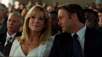 Movie still from “The Blind Side” (2009), directed by John Lee Hancock – A man and a woman sitting next to each other in front of a group of people; Close Up shot, Over the shoulder angle