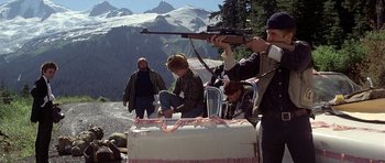 Movie still from “The Deer Hunter” (1978), directed by Michael Cimino – A group of people standing around a table with a rifle; Wide shot, High angle