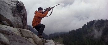 Movie still from “The Deer Hunter” (1978), directed by Michael Cimino – A man in an orange jacket is holding a rifle and kneeling on a rock in front of a forest; Wide shot, Low angle