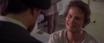 Movie still from “The Deer Hunter” (1978), directed by Michael Cimino – A man with curly brown hair and a hat talking to another man in a room; Close Up shot, Over the shoulder angle