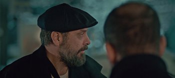 Movie still from “10 Days of a Bad Man” (2023), directed by Uluç Bayraktar – A man with a beard wearing a black hat; Close Up shot, Over the shoulder angle