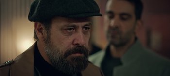 Movie still from “10 Days of a Bad Man” (2023), directed by Uluç Bayraktar – A man with a beard wearing a hat; Close Up shot, Over the shoulder angle