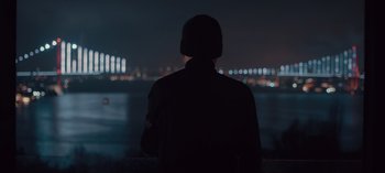 Movie still from “10 Days of a Bad Man” (2023), directed by Uluç Bayraktar – A man standing in front of a body of water at night; Medium shot, Over the shoulder angle
