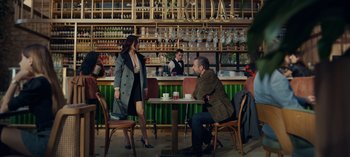 Movie still from “10 Days of a Bad Man” (2023), directed by Uluç Bayraktar – A man and a woman sitting at a table in front of a bar; Wide shot, Over the shoulder angle