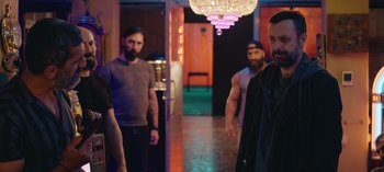 Movie still from “10 Days of a Good Man” (2023), directed by Uluç Bayraktar – A group of men standing in a room with lights in the background; Medium shot, Over the shoulder angle