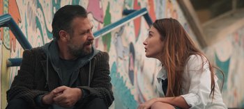 Movie still from “10 Days of a Good Man” (2023), directed by Uluç Bayraktar – A man and a woman sitting next to each other in front of a mural; Medium shot, Over the shoulder angle