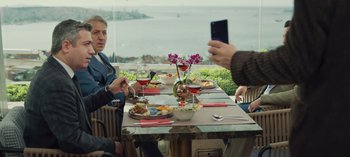 Movie still from “10 Days of a Good Man” (2023), directed by Uluç Bayraktar – A person taking a picture of a group of people sitting at a dinner table; Medium shot, Over the shoulder angle