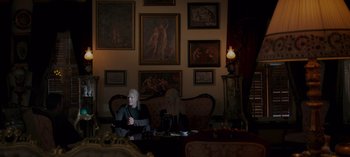 Movie still from “10 Days of a Good Man” (2023), directed by Uluç Bayraktar – A woman sitting on a couch in front of a wall of pictures; Wide shot, Over the shoulder angle