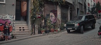 Movie still from “10 Days of a Good Man” (2023), directed by Uluç Bayraktar – A car parked on the side of the road next to a building with a mural on the side; Extreme Wide shot, Low angle
