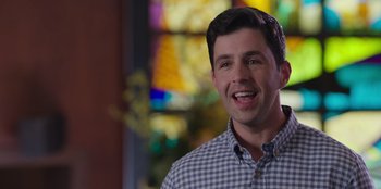 Movie still from “13: The Musical” (2022), directed by Tamra Davis – A young man smiles while standing in front of a stained glass window; Close Up shot, Over the shoulder angle