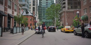 Movie still from “13: The Musical” (2022), directed by Tamra Davis – A man riding a bike down a street next to cars; Wide shot, High angle