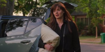 Movie still from “13: The Musical” (2022), directed by Tamra Davis – A woman holding a pillow while standing in front of a car; Medium shot, Over the shoulder angle