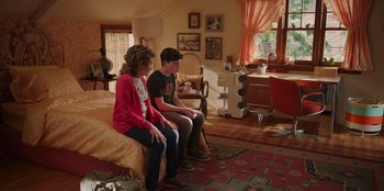 Movie still from “13: The Musical” (2022), directed by Tamra Davis – A woman and a boy sitting on a bed in a room; Wide shot, High angle