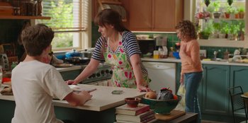 Movie still from “13: The Musical” (2022), directed by Tamra Davis – A woman in an apron standing in a kitchen; Medium shot, Over the shoulder angle