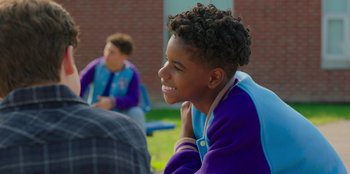 Movie still from “13: The Musical” (2022), directed by Tamra Davis – A young boy sitting on the grass in front of a brick building; Close Up shot, Over the shoulder angle