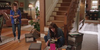 Movie still from “13: The Musical” (2022), directed by Tamra Davis – A woman sitting on the floor in front of a bunch of boxes; Wide shot, High angle