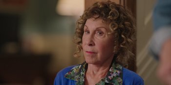 Movie still from “13: The Musical” (2022), directed by Tamra Davis – An older woman with curly blonde hair wearing a blue shirt; Close Up shot, Over the shoulder angle