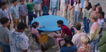 Movie still from “13: The Musical” (2022), directed by Tamra Davis – A group of people gathered around a blue table; Wide shot, High angle