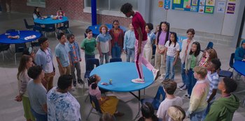 Movie still from “13: The Musical” (2022), directed by Tamra Davis – A group of people standing around a round table; Extreme Wide shot, Over the shoulder angle