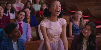 Movie still from “13: The Musical” (2022), directed by Tamra Davis – A girl in a pink dress is laughing in front of a group of people; Medium shot, Over the shoulder angle