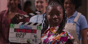 Movie still from “13: The Musical” (2022), directed by Tamra Davis – A woman is holding a clapperboard in front of a crowd; Medium shot, Over the shoulder angle
